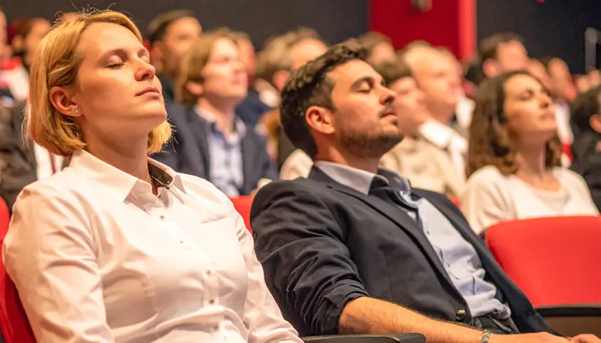 Deelnemers in een congreszaal met rode stoelen die met gesloten ogen een korte stilte- of ademhalingsoefening ervaren tijdens de Full Day Experience.